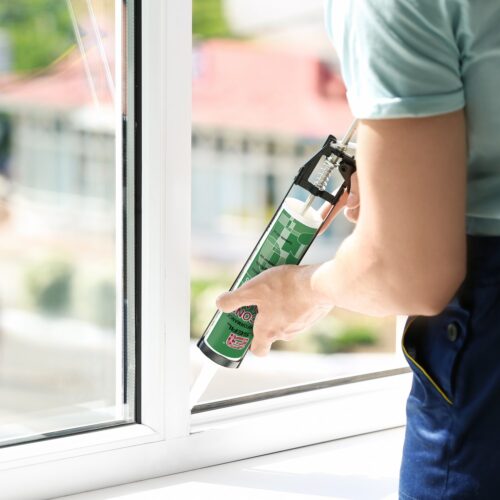 A person wearing work clothes stands by a window, holding a tube of silicone sealant, sealing the gaps.